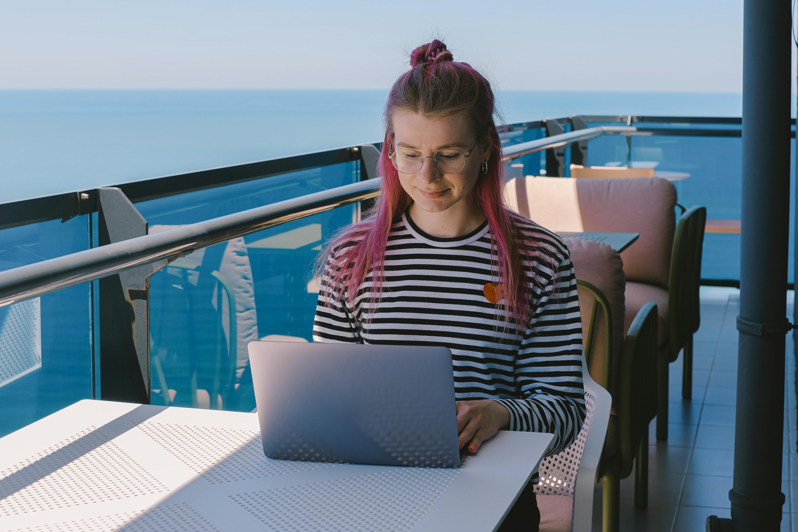 Young woman freelancing with a laptop on a sunny oceanfront balcony, embracing remote work lifestyle.