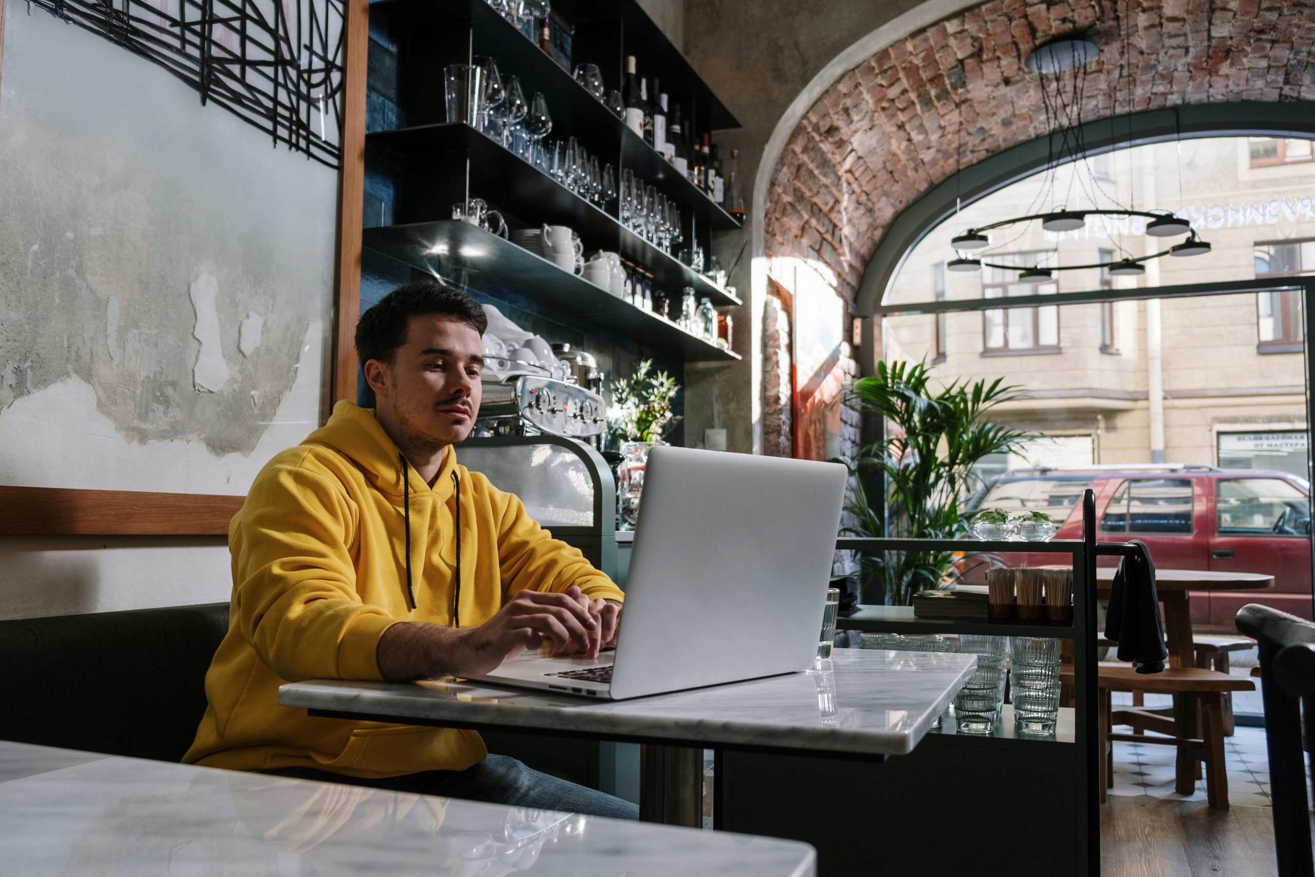 A freelancer in a yellow hoodie works on a laptop in a trendy brick-arched café.