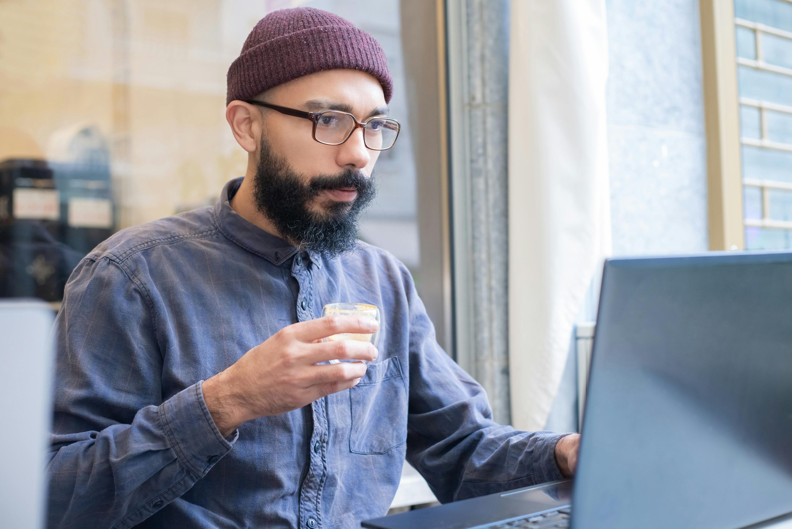 Asian man with eyeglasses working on laptop in a café while holding a coffee cup.