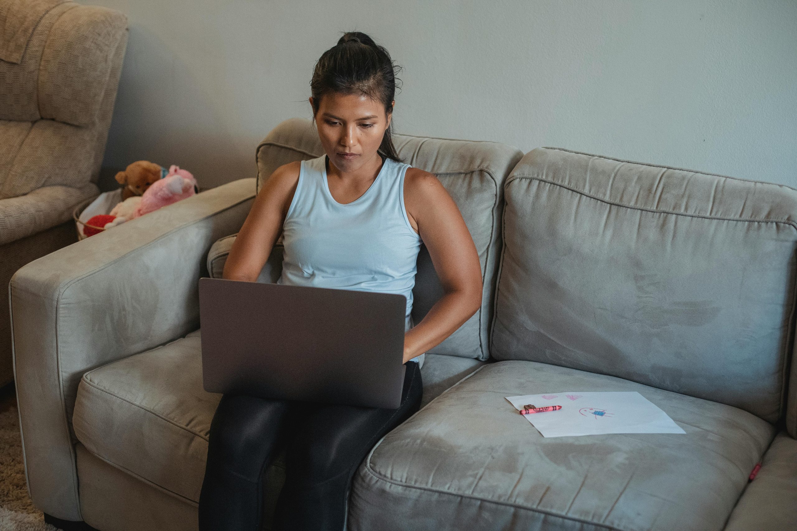 Adult woman working remotely on netbook while sitting on sofa near child drawing at home