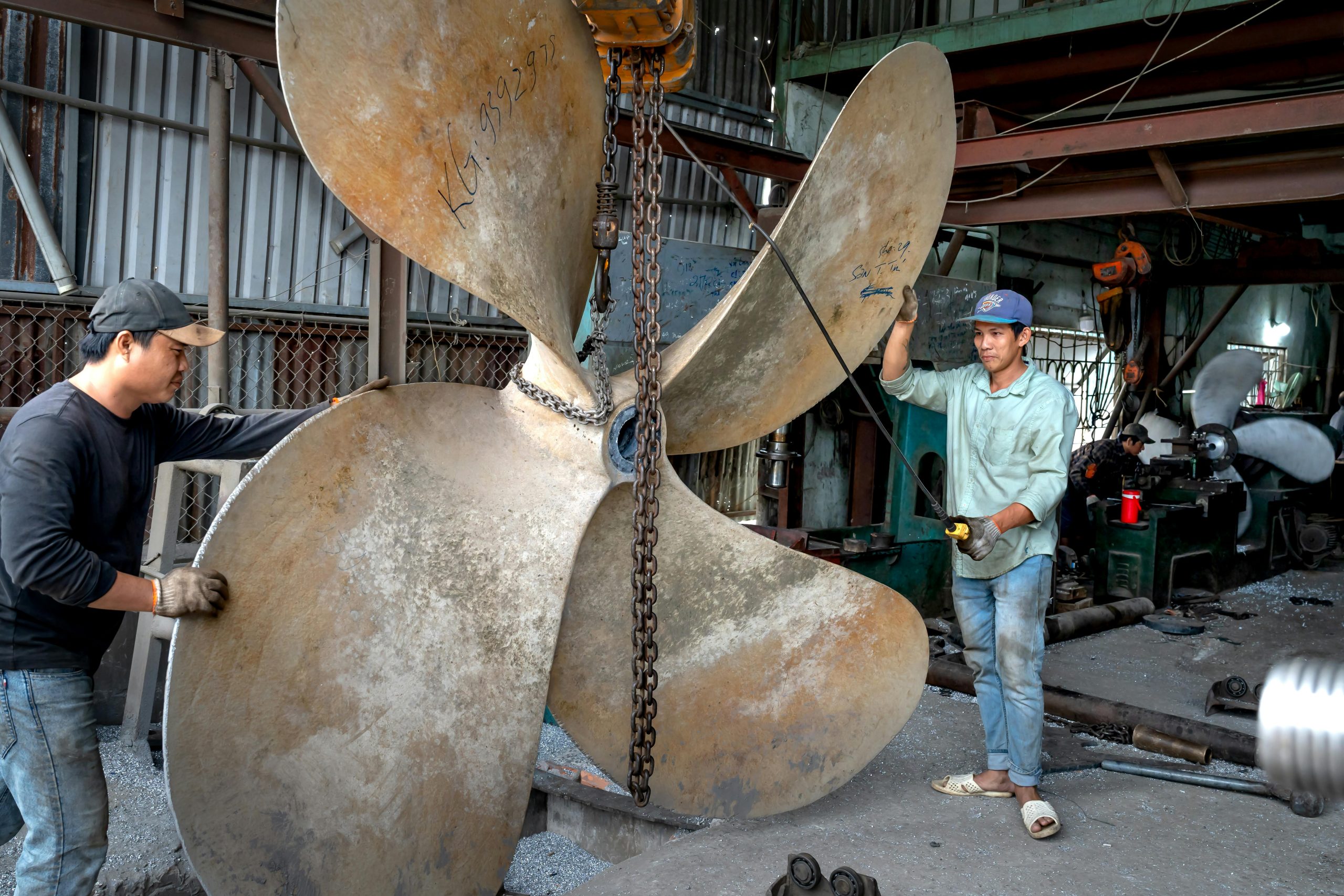 Two men assembling a large metal ship propeller in an industrial workshop setting.