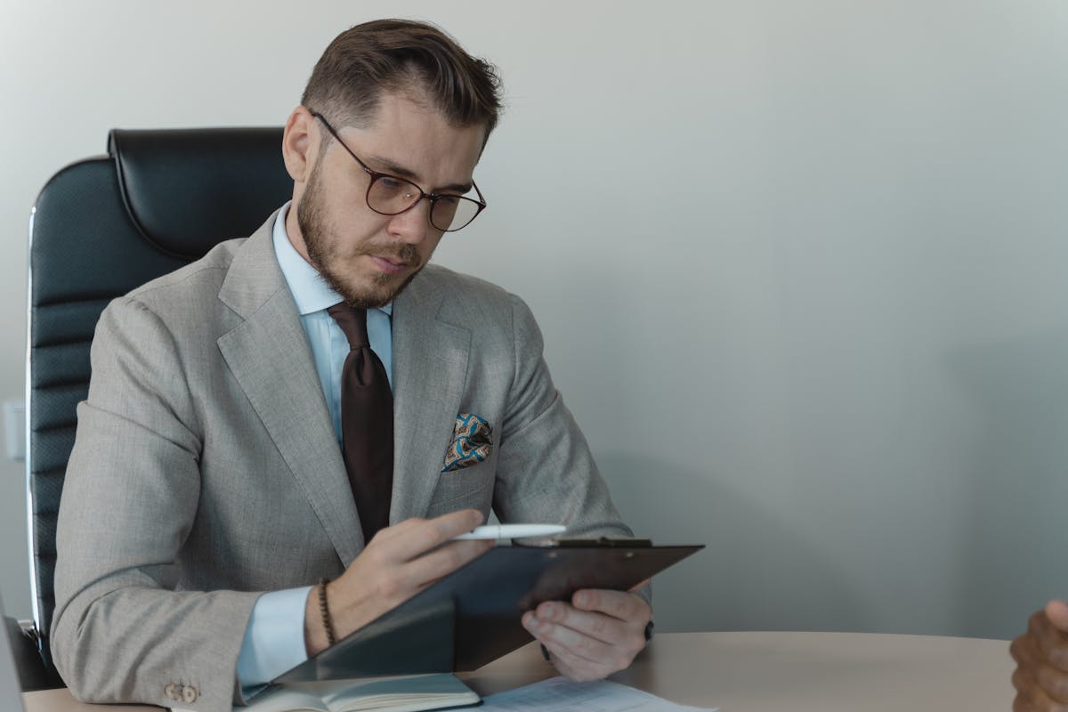 Businessman in a gray suit with glasses reviewing documents on a clipboard in an office.